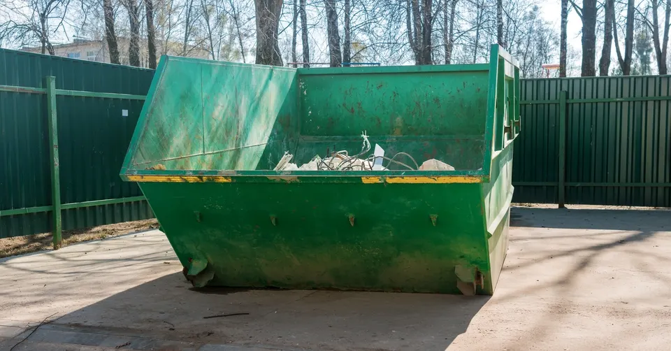 A large green dumpster on a concrete slab in front of a green solid metal fence.