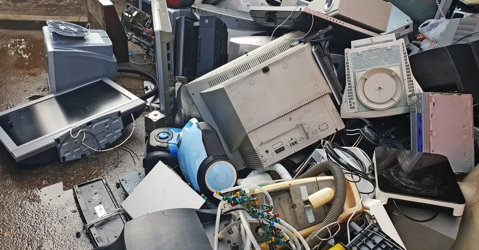 A pile of old electronics on the ground. The pile includes TVs, desktop computers, and other miscellaneous items.