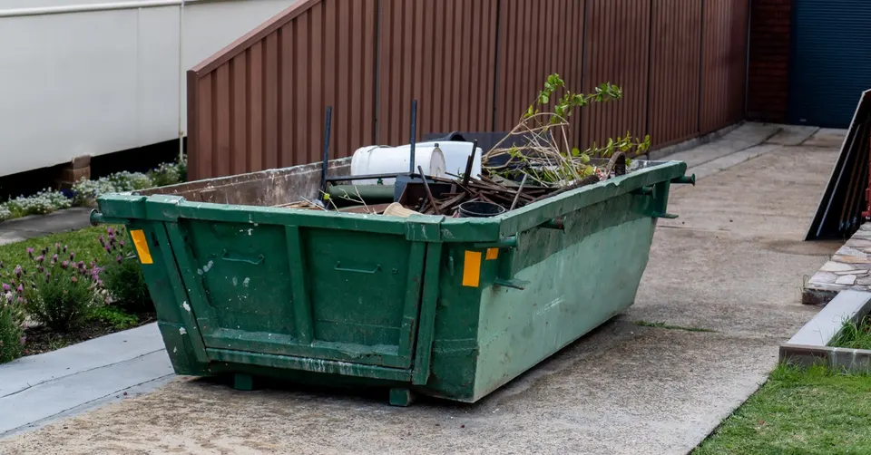 A large dumpster sits in a driveway. The dumpster holds yard and home debris, including tree branches and paint containers.