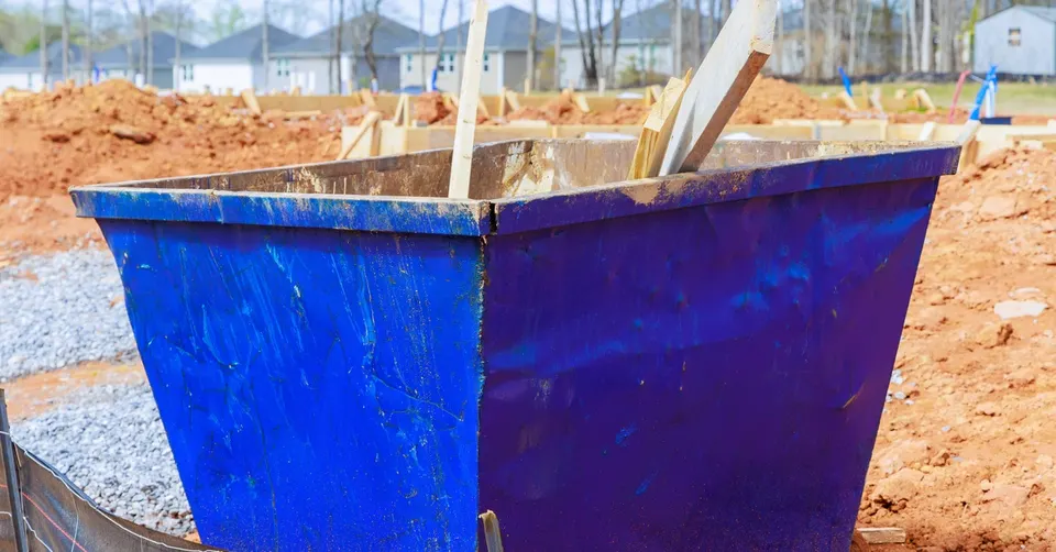 A blue dumpster sits on a construction site near dirt piles. The dumpster has wood planks sticking out of it.