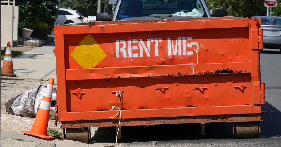 A close-up of an orange dumpster sitting on a curb. It says "RENT ME" in white spray-painted letters.