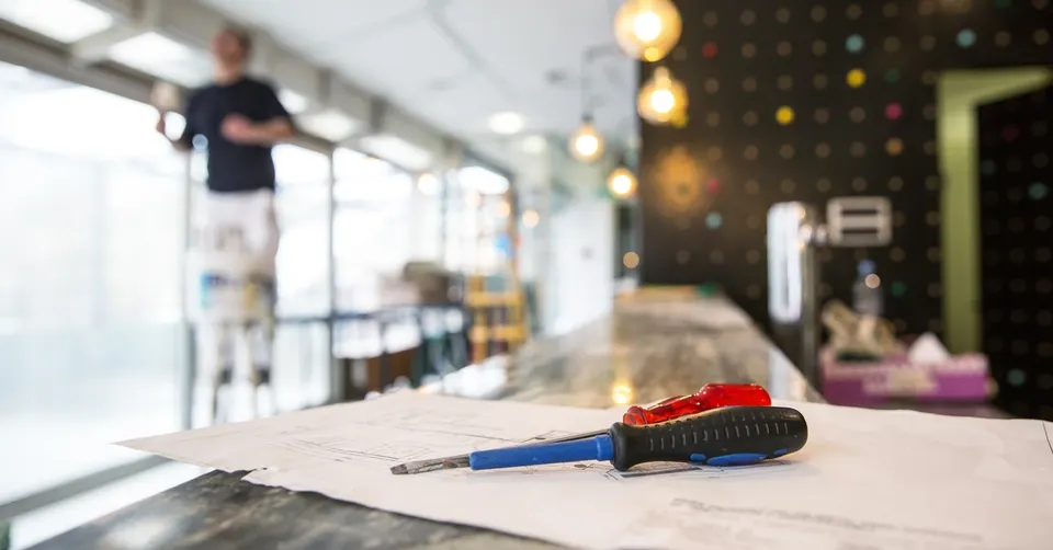 Two screwdrivers are sitting on a table in the foreground. In the background, there is a person out of focus, standing on a ladder.
